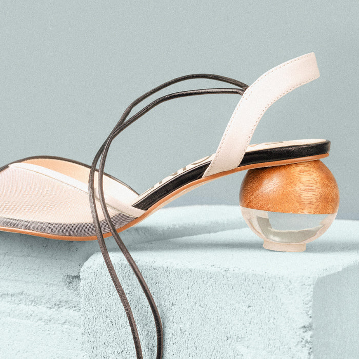 Stylish designer sandal with a unique wooden and glass ball heel, set against a soft blue background.