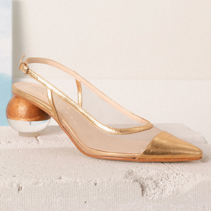 A gold slingback shoe with a pointed toe and unique round heel, featuring a transparent mesh upper set on a cement block against a white background