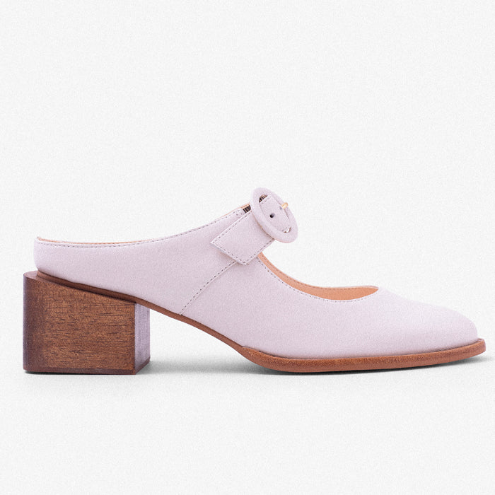 Light purple mules with a square, wooden heel and a round buckle detail, designed for stylish comfort, set against a solid powder blue background.