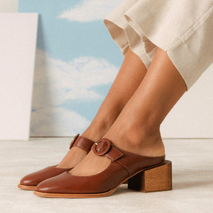 A close-up of a woman's feet wearing brown leather mules with a round buckle detail and a square wooden, paired with light beige trousers against a sky background.