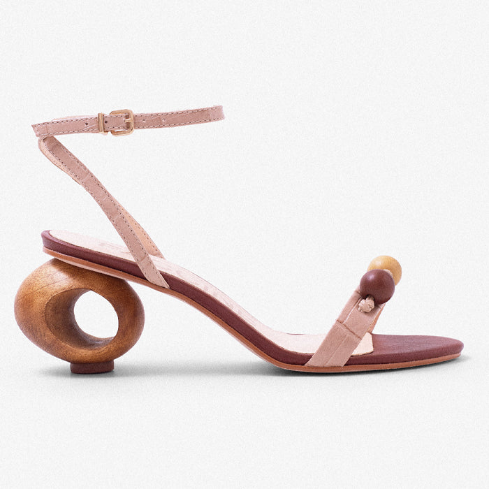 Stylish nude leather sandal with a curved wooden heel and decorative wooden beads on the strap, set against a soft blue sky background.