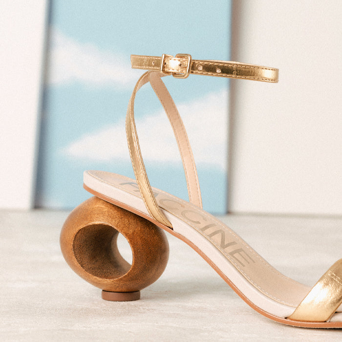 Stylish gold leather strap sandal with a curved wooden heel set against a soft blue sky background.