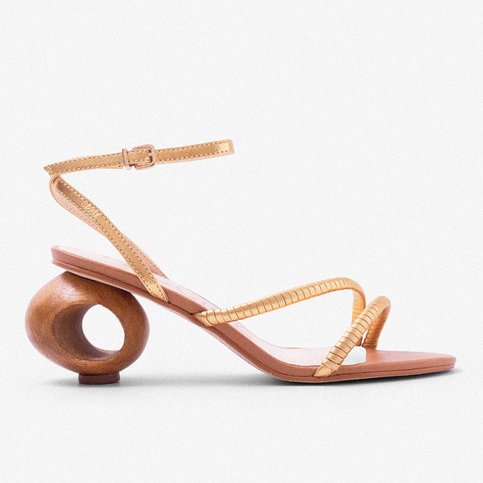 Gold strappy sandal with unique wooden circular heel, showcasing a stylish blend of elegance and contemporary design set against a solid powder blue background