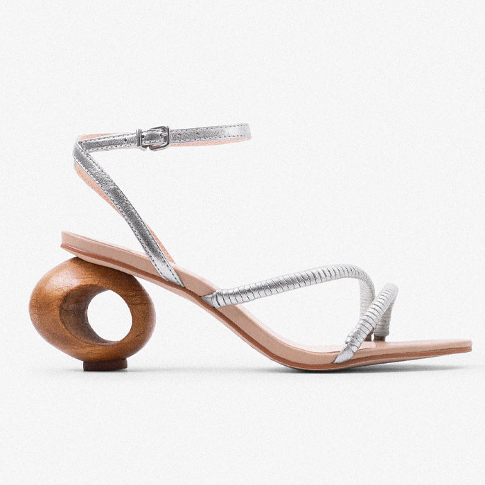 Silver strappy sandal with unique wooden circular heel, showcasing a stylish blend of elegance and contemporary design set against a solid powder blue background