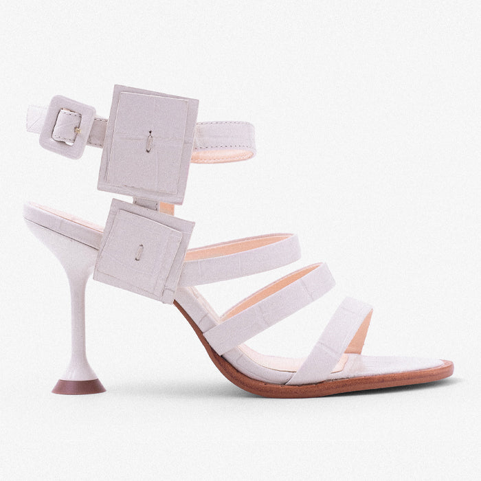 Stylish white high-heeled sandal with geometric buckle detail and multiple straps, set against a solid powder blue background.