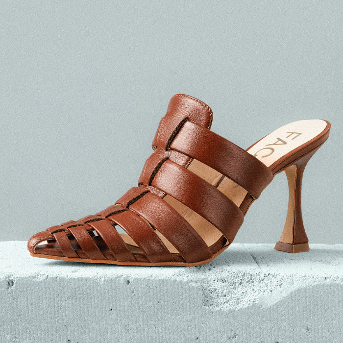 A stylish brown leather heeled sandal with elegant crisscross straps and a slender stiletto heel, displayed on a powder blue concrete surface.