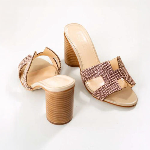 A pair of stylish beige heeled mules with a sparkling rose gold beads embellished on a strap, featuring a chunky wooden heel and an H-shape open toe design.