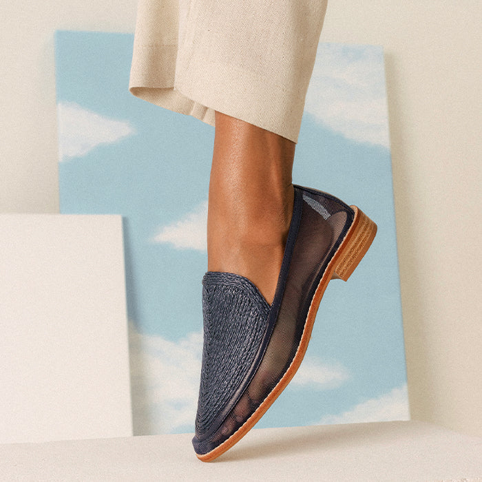 A close-up of a woman's foot wearing a navy slip-on shoe with mesh detailing, and a wooden heel, set against a soft blue sky background.